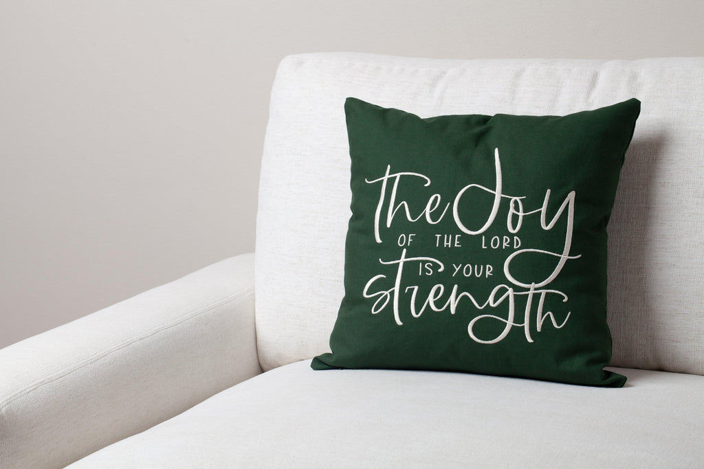 Green Joy & Strength embroidered throw pillow on white sofa – vibrant Nehemiah 8:10 verse uplifts contemporary faith-inspired space